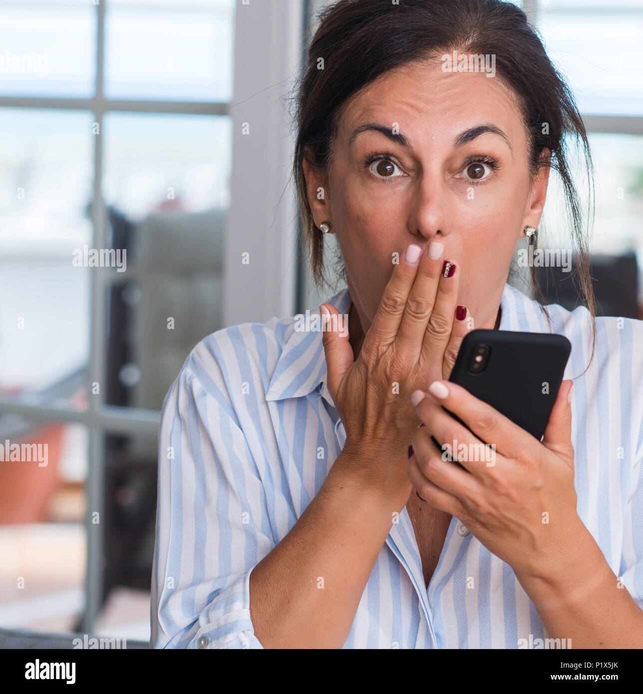 Middle aged woman using smartphone cover mouth with hand shocked with ...