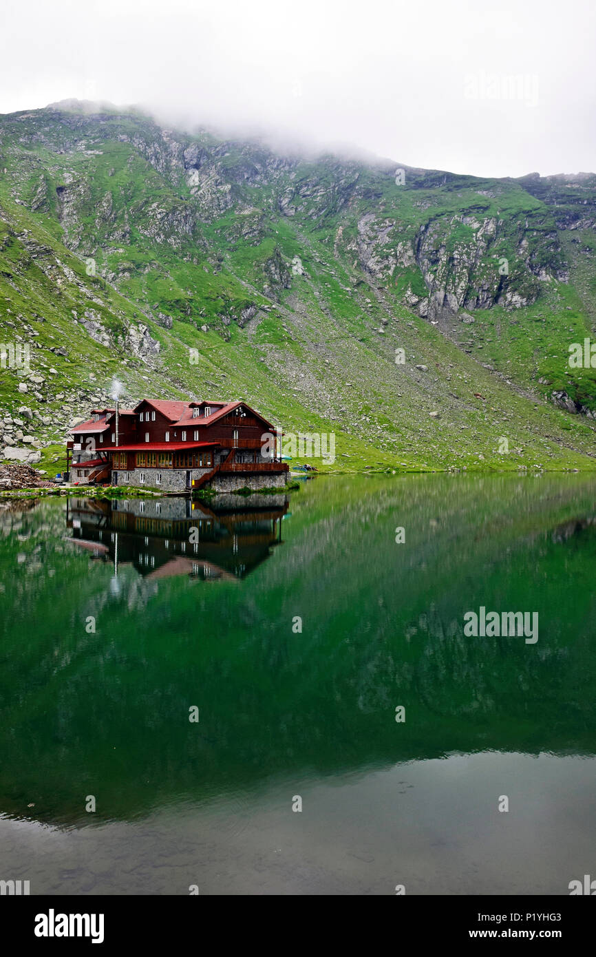 Chalet and alpine steppe covered, clouded rocky hills reflected in the mirror-like surface of Bâlea lake , Southern Carpathians Stock Photo