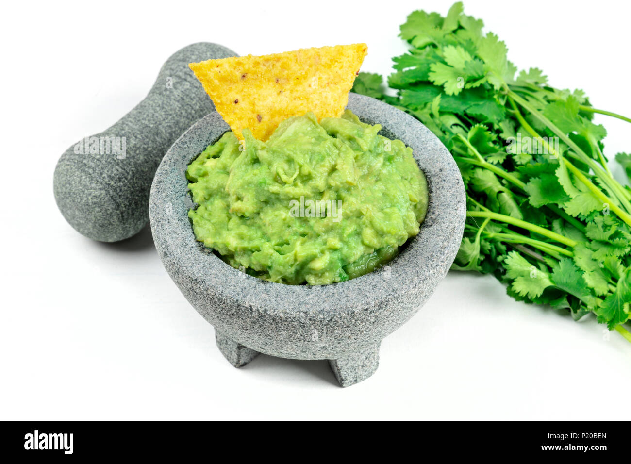 A photo of guacamole sauce in a molcajete, traditional Mexican mortar, on a white background, with a pestle. a bunch of cilantro, a nacho chip, and a  Stock Photo