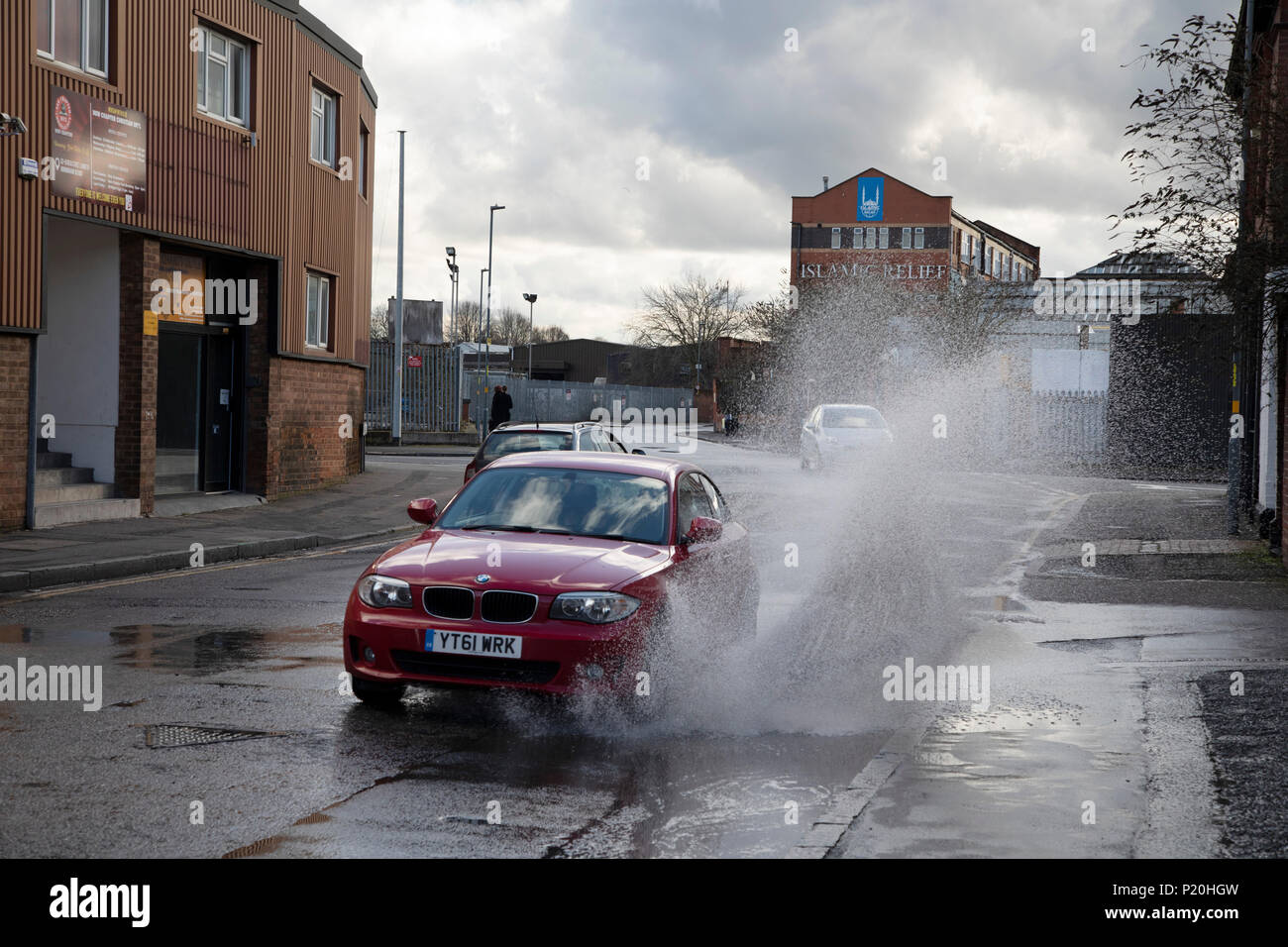 Car drives through a large puddle near to Islamic Relief Worldwide in Digbeth, Birmingham, England, United Kingdom. Digbeth is an area of Central Birmingham, England. Islamic Relief Worldwide is an international humanitarian organisation that provides development programs and humanitarian relief around the globe, regardless of race, political affiliation, gender or belief. Following the destruction of the Inner Ring Road, Digbeth is now considered a district within Birmingham City Centre. As part of the Big City Plan, Digbeth is undergoing a large redevelopment scheme that will regenerate the  Stock Photo