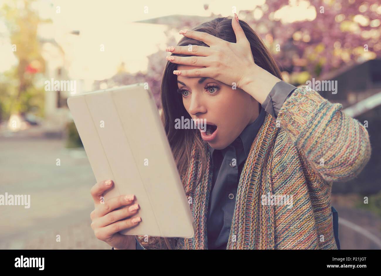 Upset woman astonished surprised by what she sees on her tablet pad standing outside in the city street Stock Photo