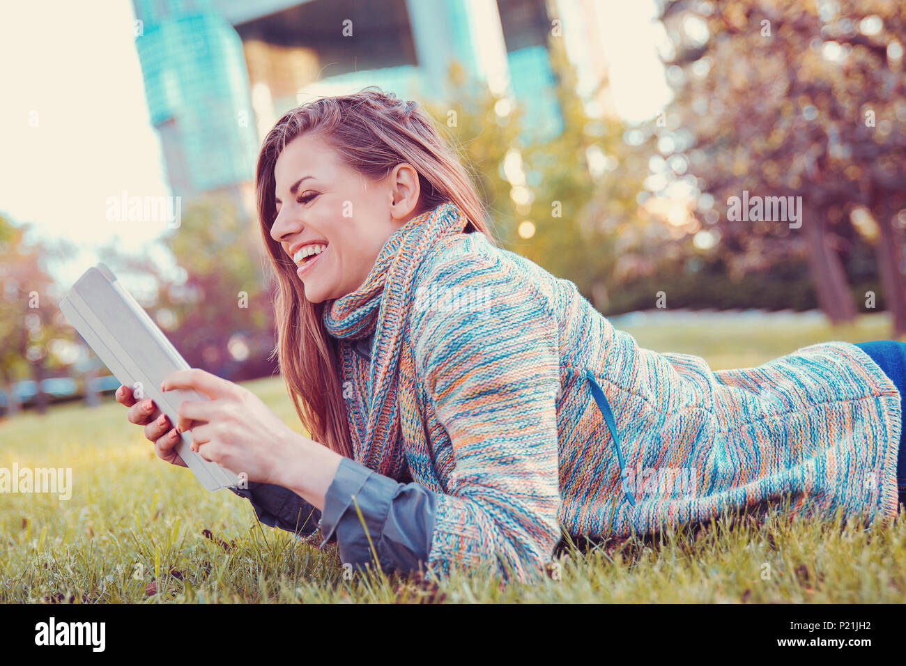 Young woman using tablet computer outdoors laying on grass in a city park, smiling. Stock Photo