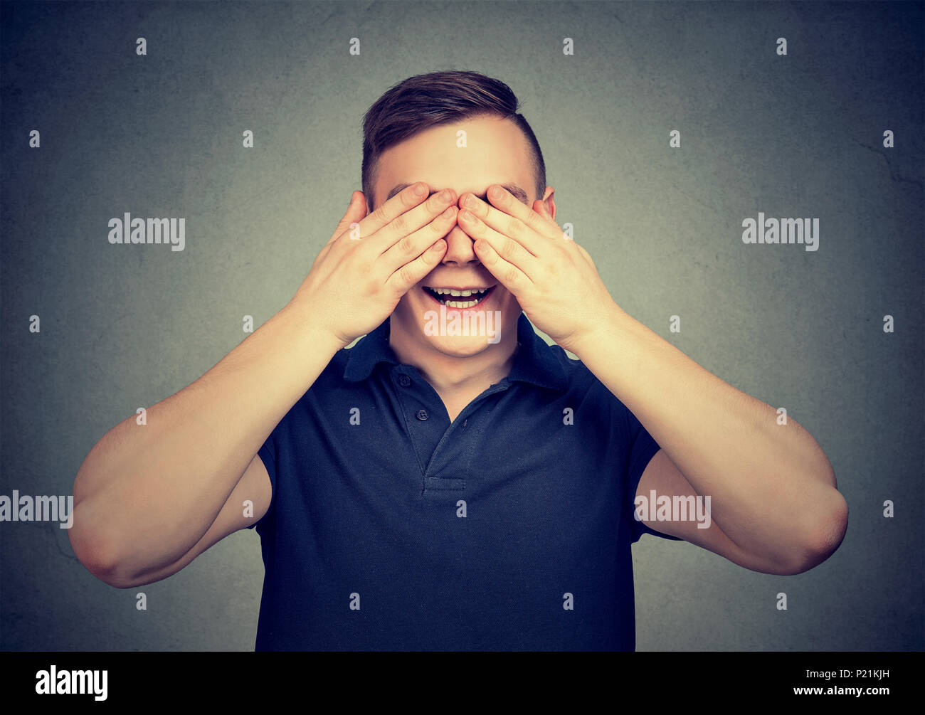 Young cheerful man covering eyes with hands anticipating surprise while posing on gray backdrop. Stock Photo