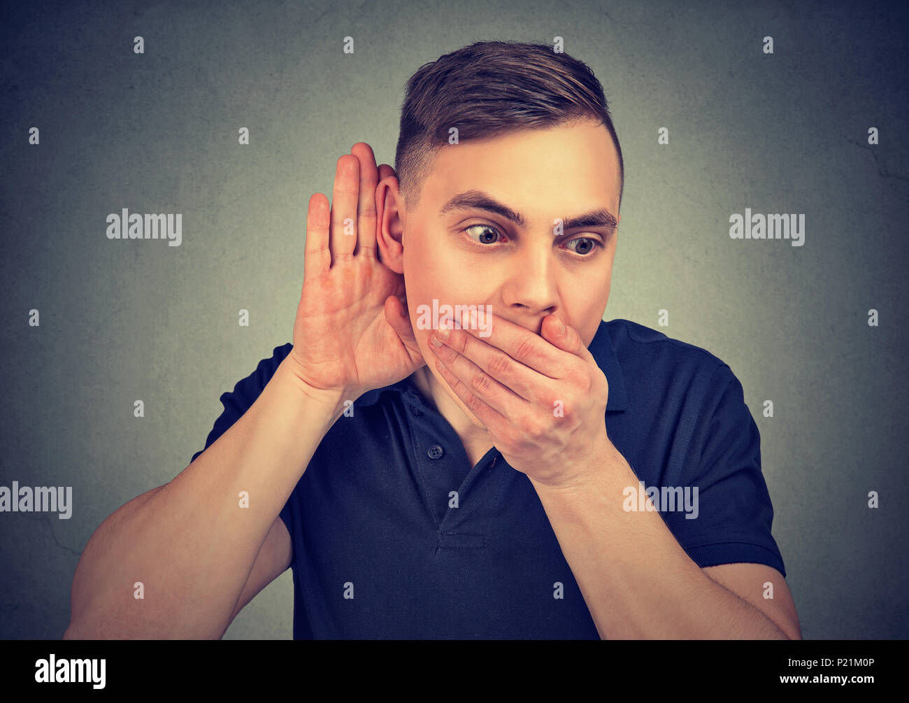 Young man holding hand near ear listening to gossips while covering mouth from amazement Stock Photo