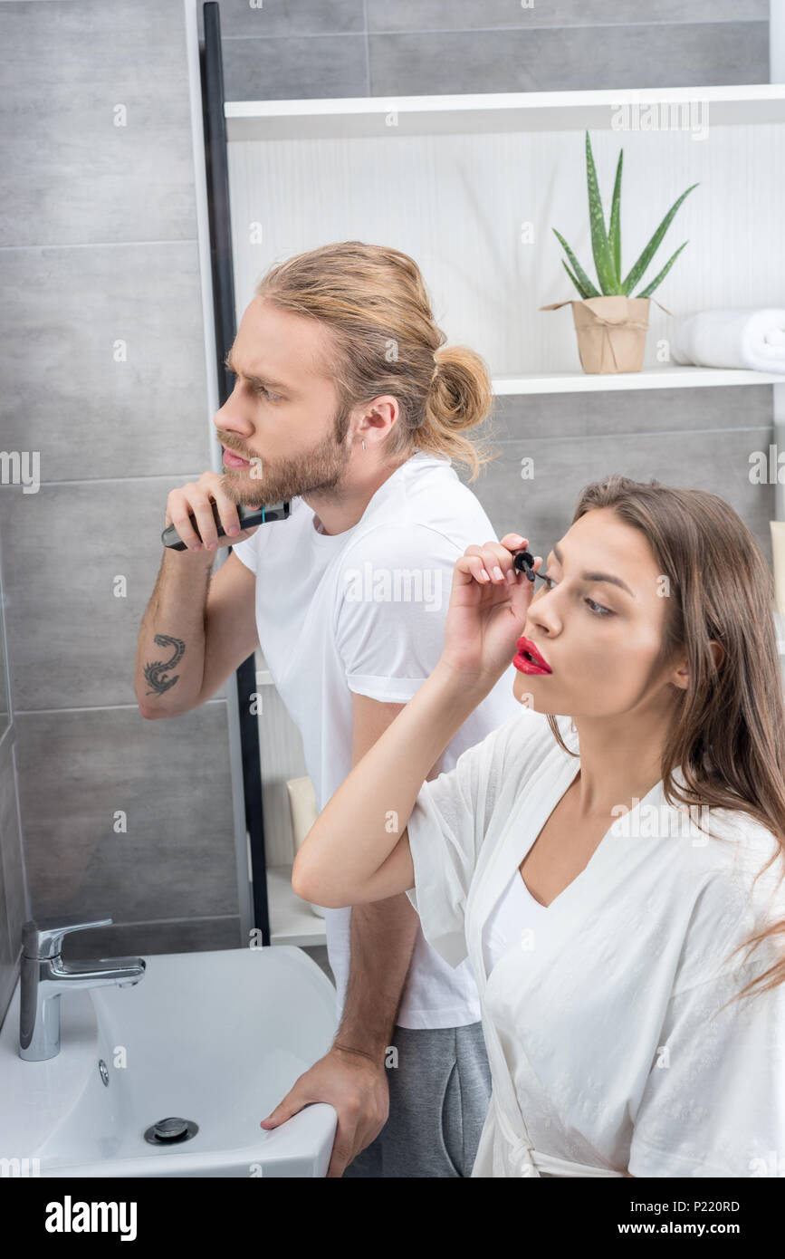 man shaving his beard while woman applying mascara in bathroom in the