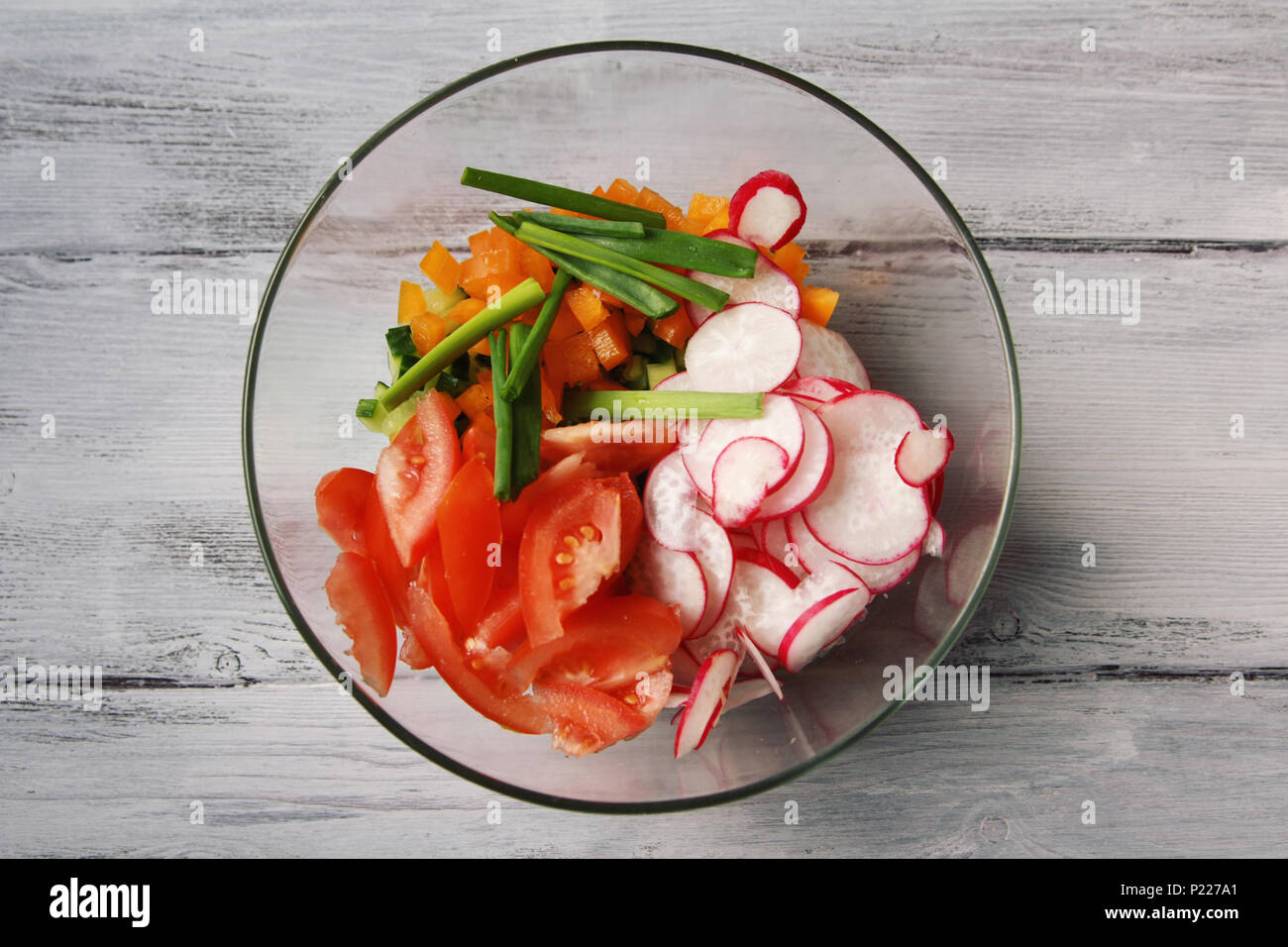Glass bowl with vegetables for a vegetarian salad. Radishes, tomatoes, celery, bell pepper, onion and cucumber. White wooden kitchen table. Close up.  Stock Photo