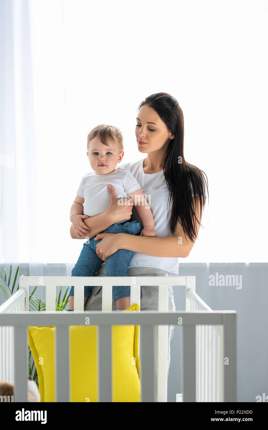 Portrait Of Mother With Little Baby In Hands Standing At Baby Crib