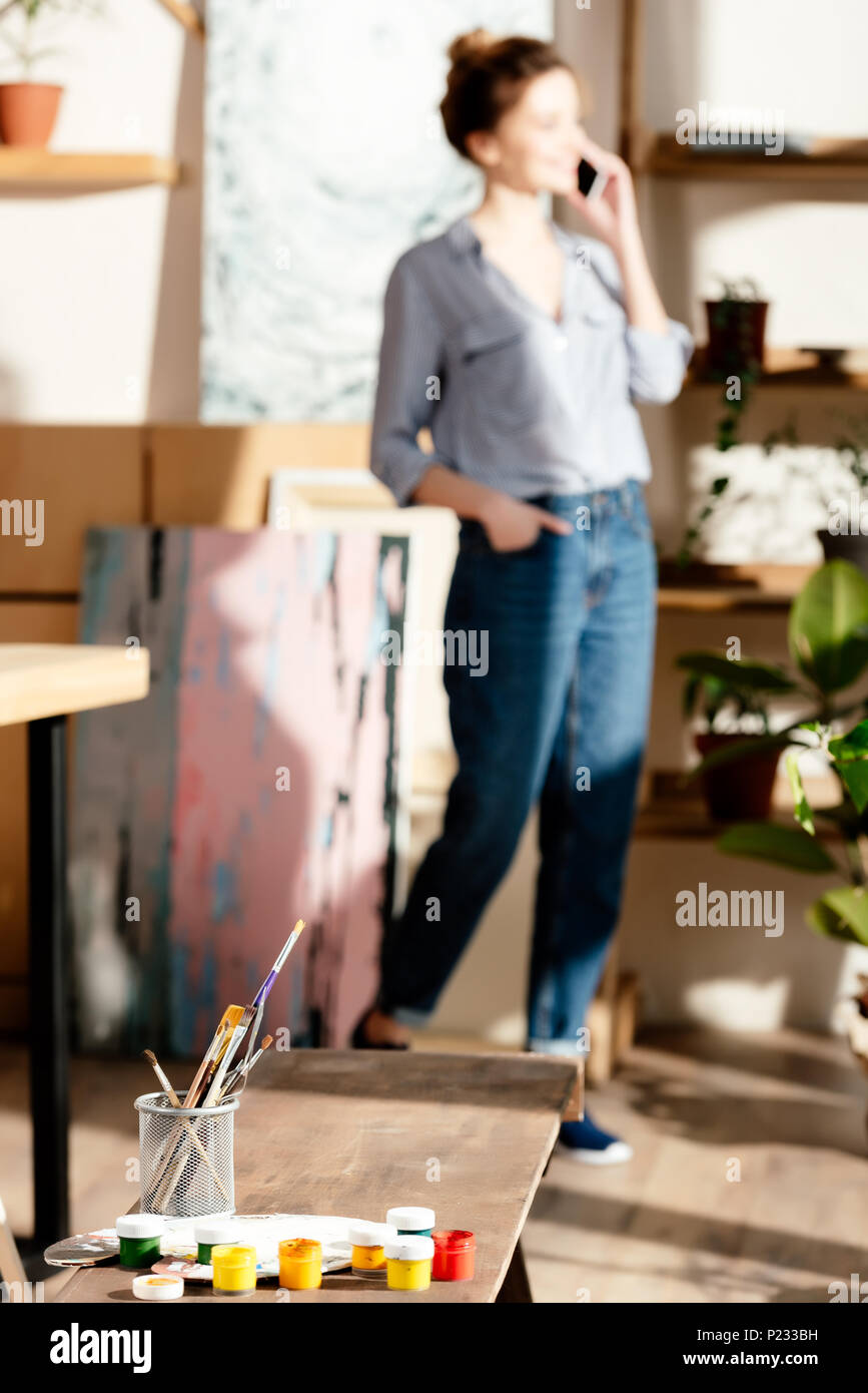 painting supplies on bench and female artist talking on smartphone behind Stock Photo