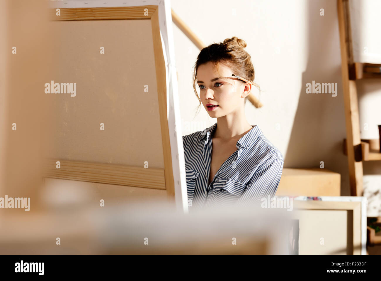 young female artist with paintbrush behind ear looking at painting Stock Photo