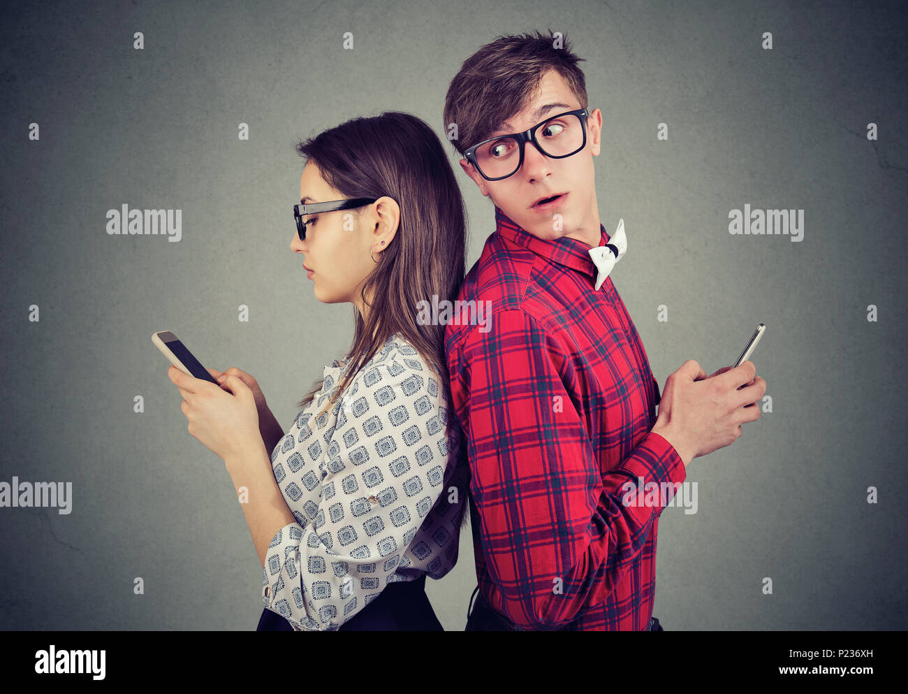 Young couple standing back to back, holding smartphones. Jealous man looking over his shoulder at his girlfriend phone trying to see what she is texti Stock Photo