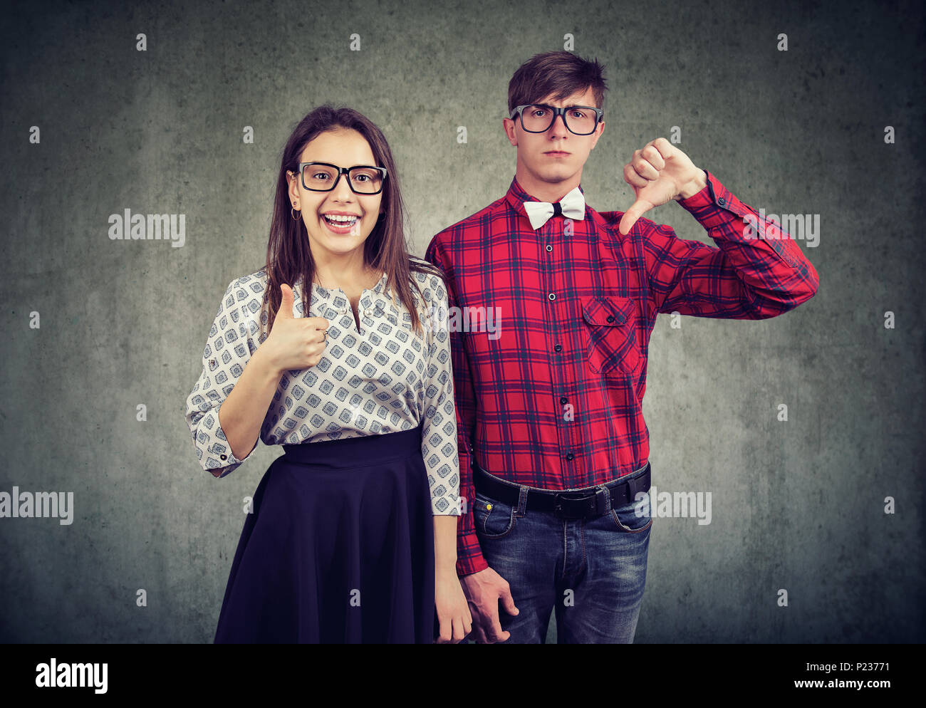 Young stylish man with thumb down standing with optimistic girl showing thumb up looking at camera. Stock Photo