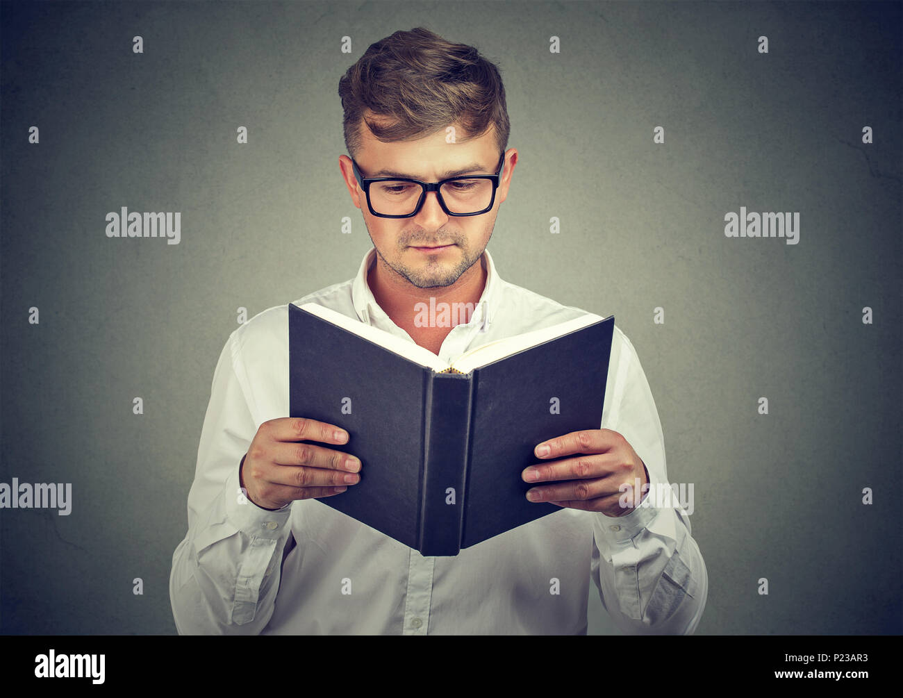 Casual young man wearing eyeglasses and reading big book learning material and looking serious. Stock Photo