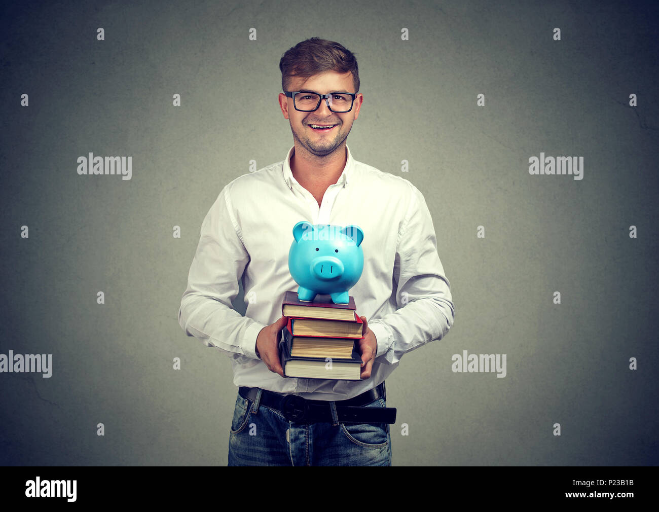 Young handsome man in eyeglasses holding stack of books and moneybox with money for university. Stock Photo