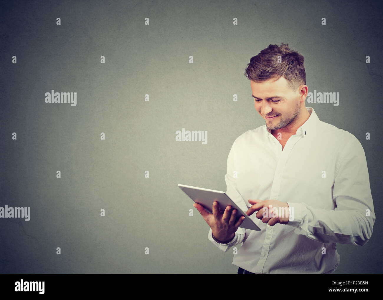Content casual guy in white shirt browsing modern tablet enjoying new device standing on gray. Stock Photo