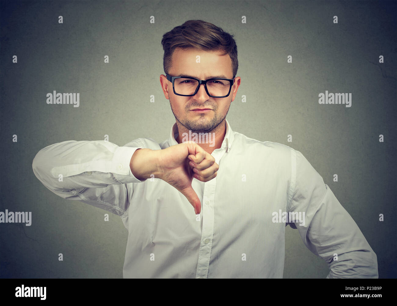 Handsome man in eyeglasses and white shirt giving opinion with thumb dump looking grumpy at camera. Stock Photo