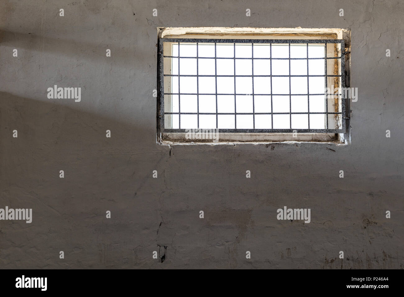 Old white wall with window covered by iron grate. Sun shining  through grill. Prison room interior . Enlightment concept. Copyspace. Stock Photo