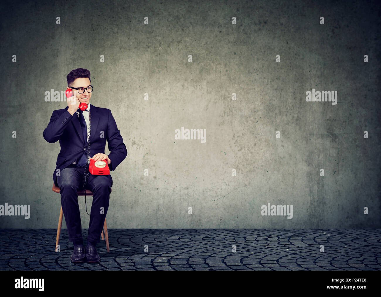 Young stylish business man sitting on chair on gray wall background holding red old fashioned telephone and talking cheerfully. Stock Photo
