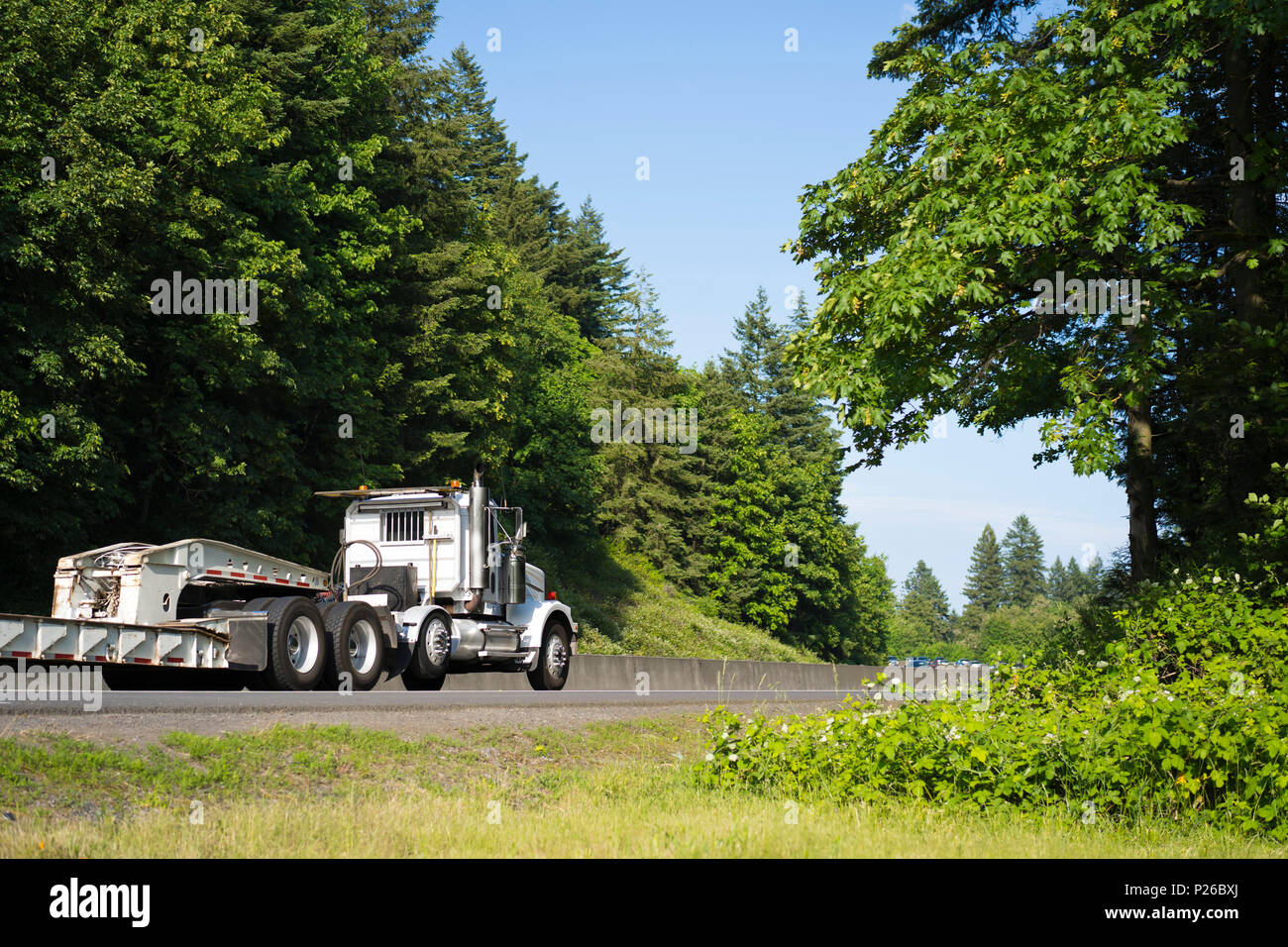 Commercial powerful big rig day cab local haul white semi truck transporting step down semi trailer on the green winding highway with trees for delive Stock Photo