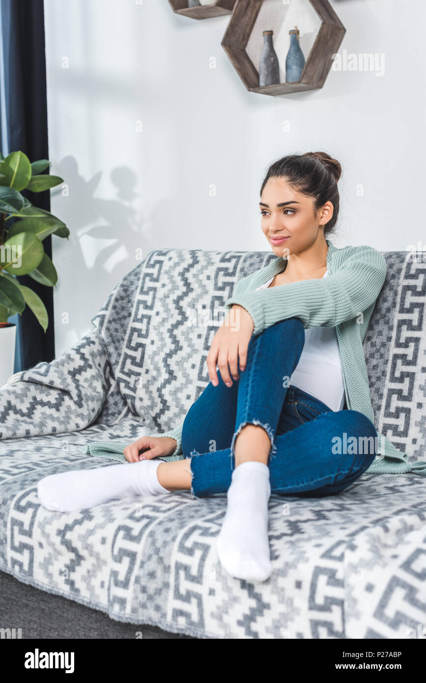 beautiful smiling young woman sitting on couch and looking away at home ...