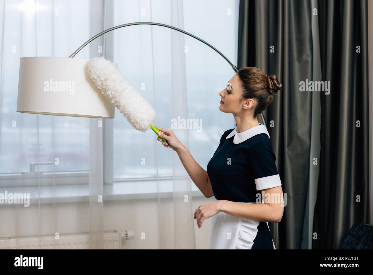 young happy maid in uniform cleaning torchere with duster Stock Photo