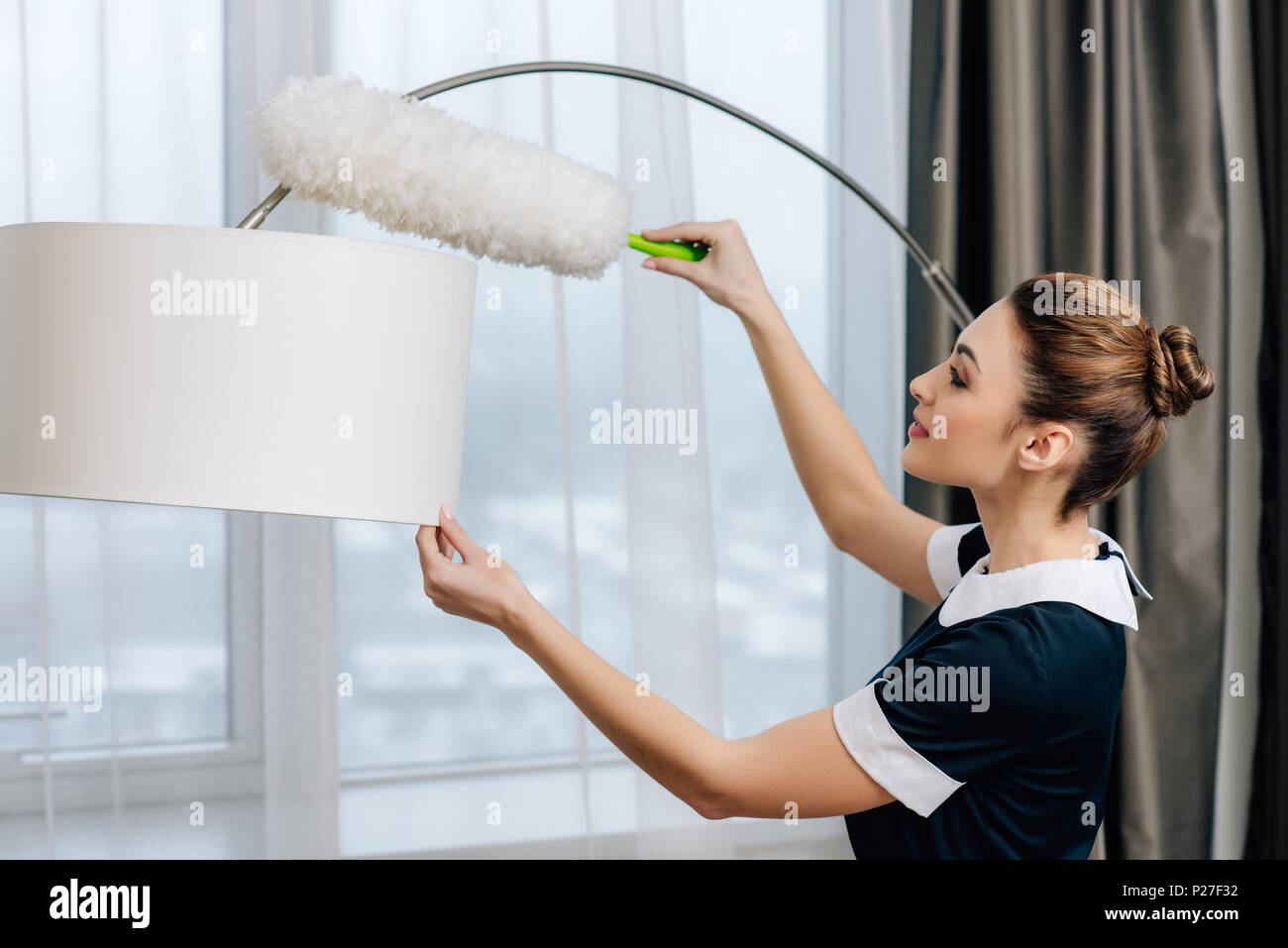 young beautiful maid in uniform cleaning torchere with duster Stock Photo