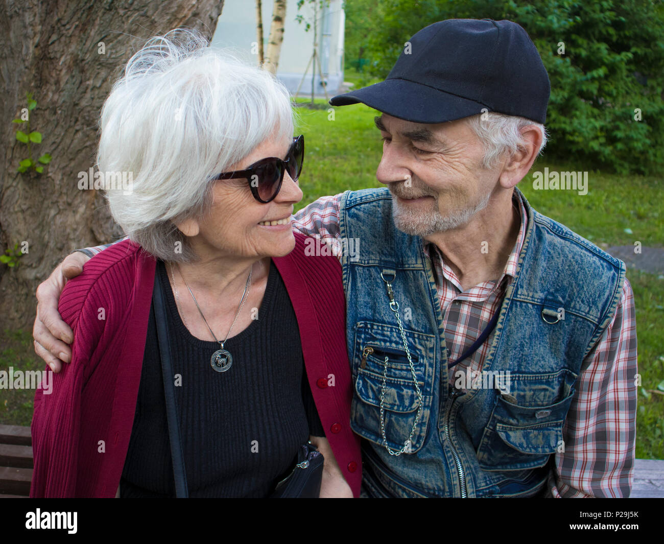 Happy elderly smiling couple sitting on the bench in the park, man looks at woman with love Stock Photo