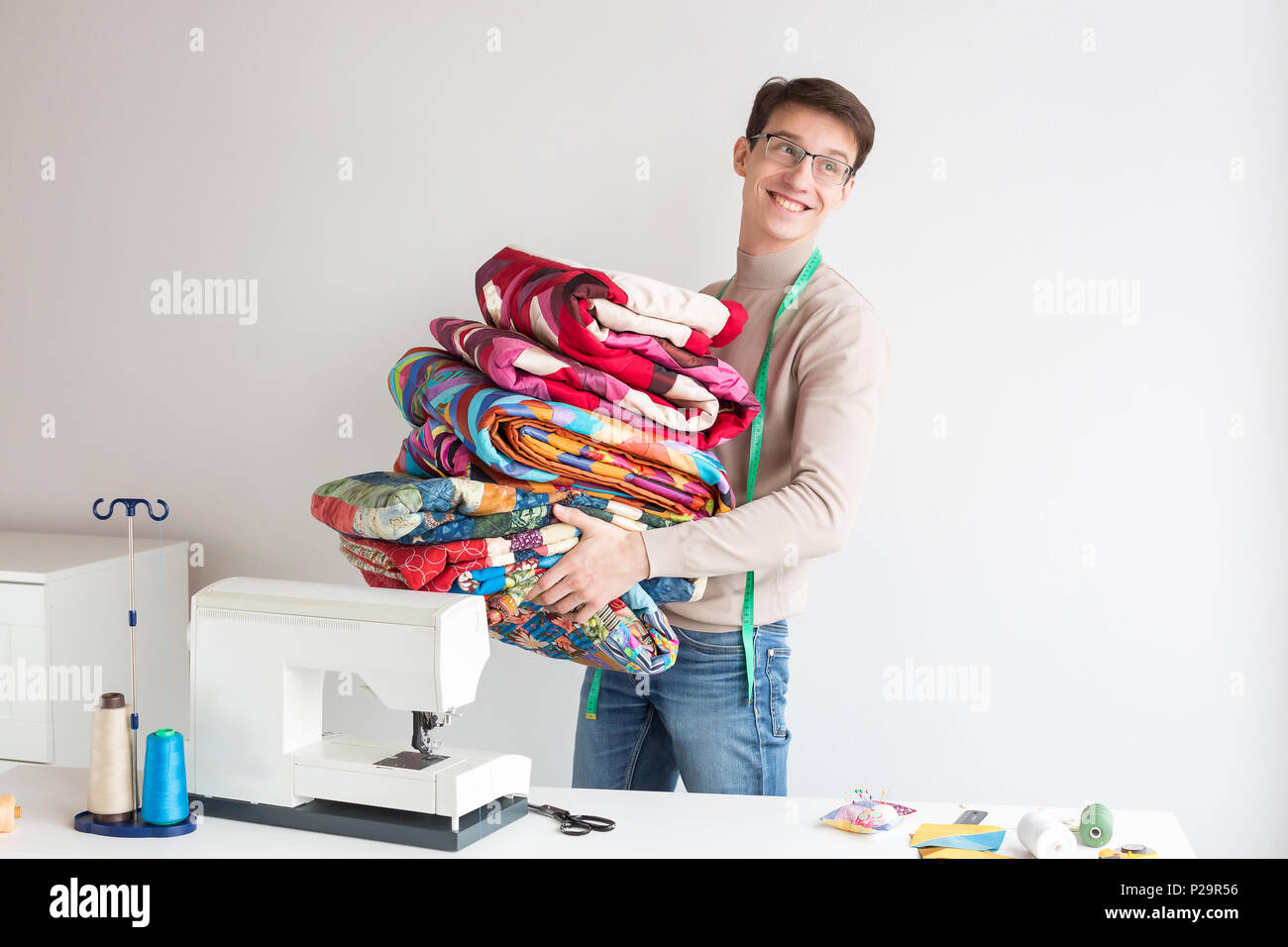 profession, business, hobby concept. there is a young tailor, who is standing in the light studio of desighn and sewing, he is holding lots of quilts  Stock Photo