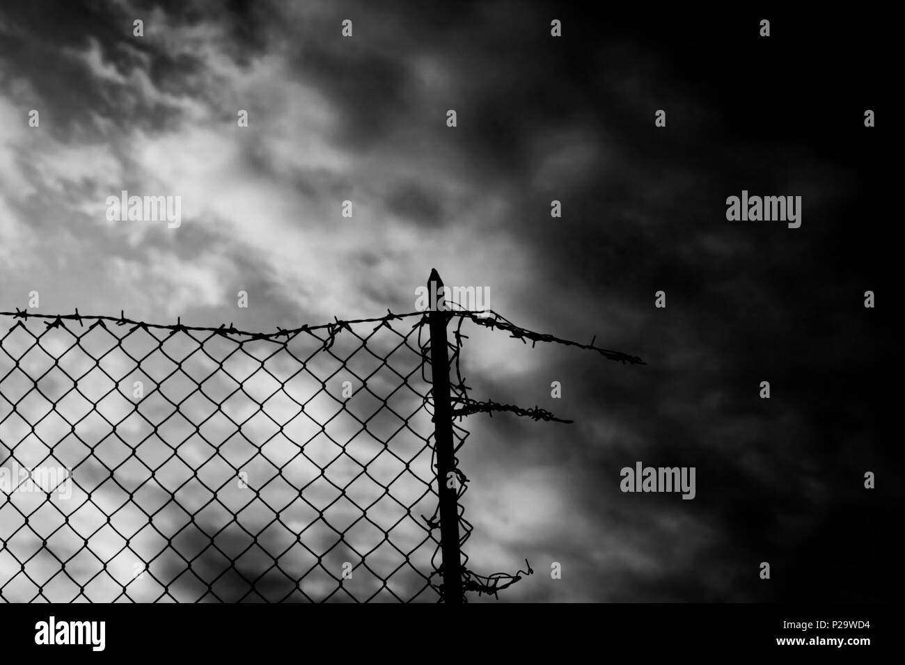 A broken fence over a dark sky. Barbed wire on top. Black-and-white shot. Symbolic shot: escapism, jail, hope, war. Stock Photo