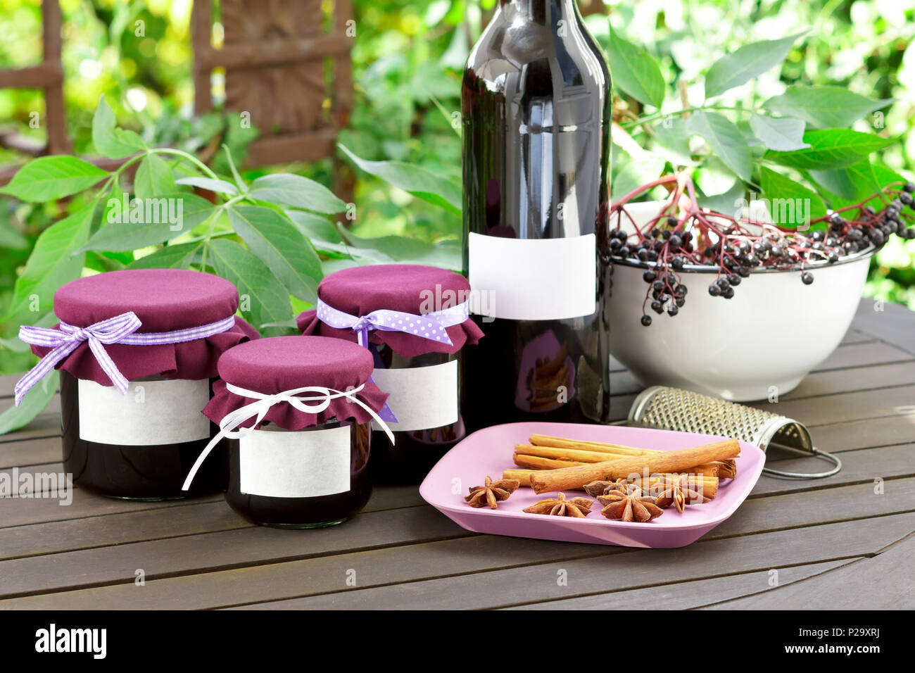 Homemade elderberry jelly and juice in a bottle and glass jars with elderberries on a wooden table outdoors in front of green leaves, copy space. Stock Photo