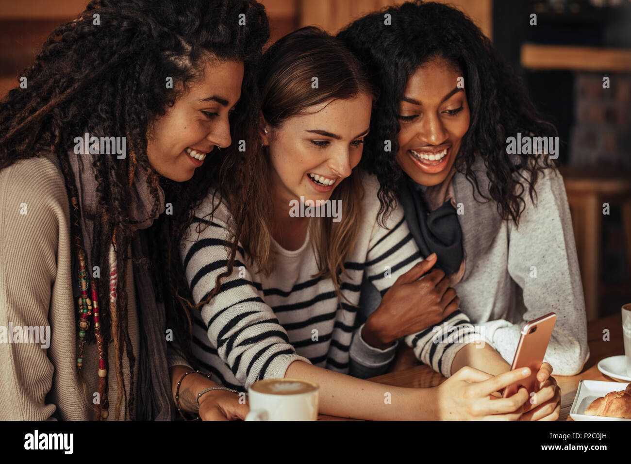 Three women sitting in a restaurant looking at mobile phone and laughing. Friends sitting at a cafe with coffee and snacks on the table looking at a m Stock Photo