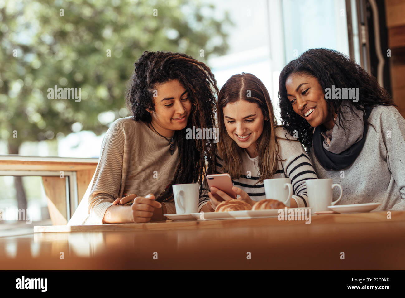 Three women sitting in a restaurant looking at mobile phone and smiling. Friends sitting at a cafe with coffee and snacks on the table looking at a mo Stock Photo