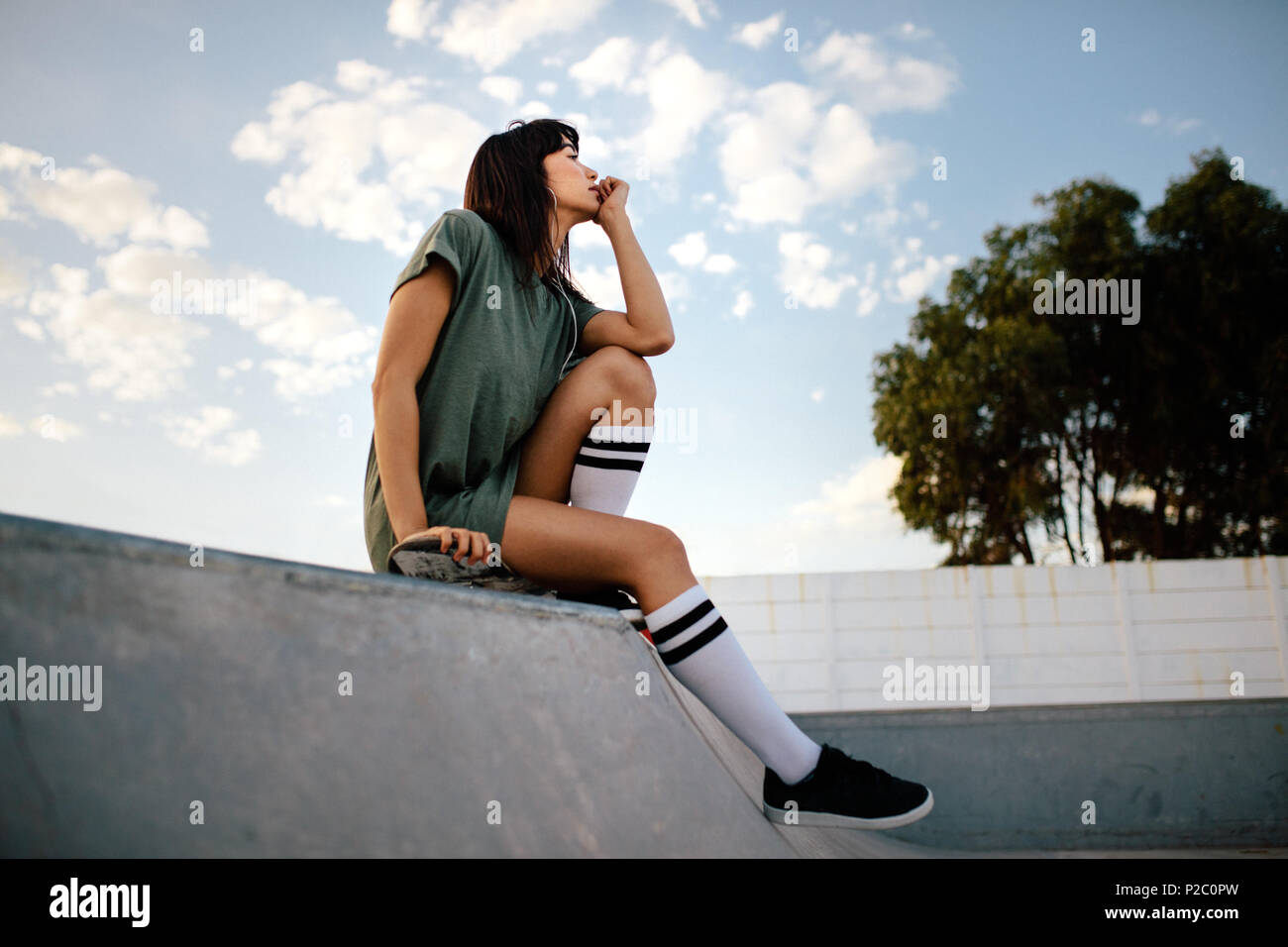 Woman sitting on skating ramp looking away and thinking at the skate park. Young female skateboarded relaxing at skate park. Stock Photo