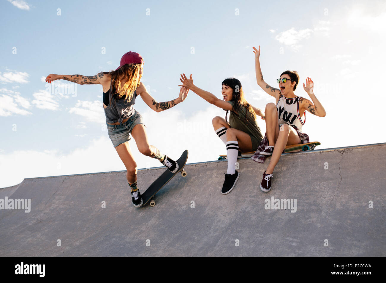 Female skateboarder riding skateboard at skate park with friends sitting on  ramp having fun. Woman skater giving high five to female friend sitting on  Stock Photo - Alamy