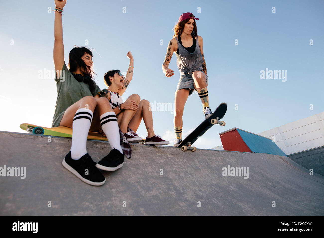 Three young female friends at skate park. Skater girl starting her routine with women friends cheering sitting on ramp. Stock Photo