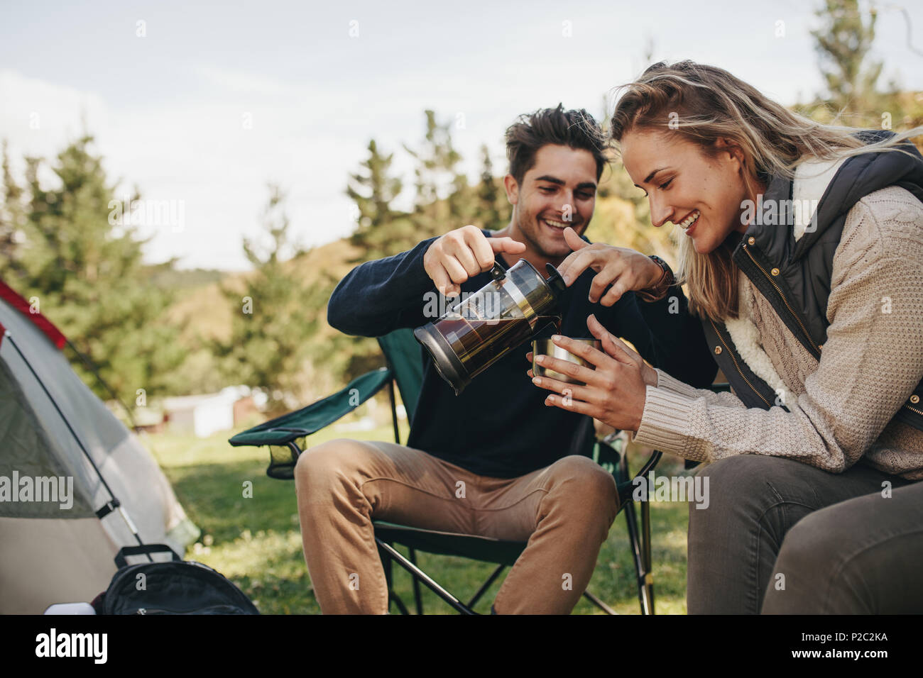 Beautiful young couple having coffee while camping in nature. Man pouring coffee in woman's cup. Stock Photo