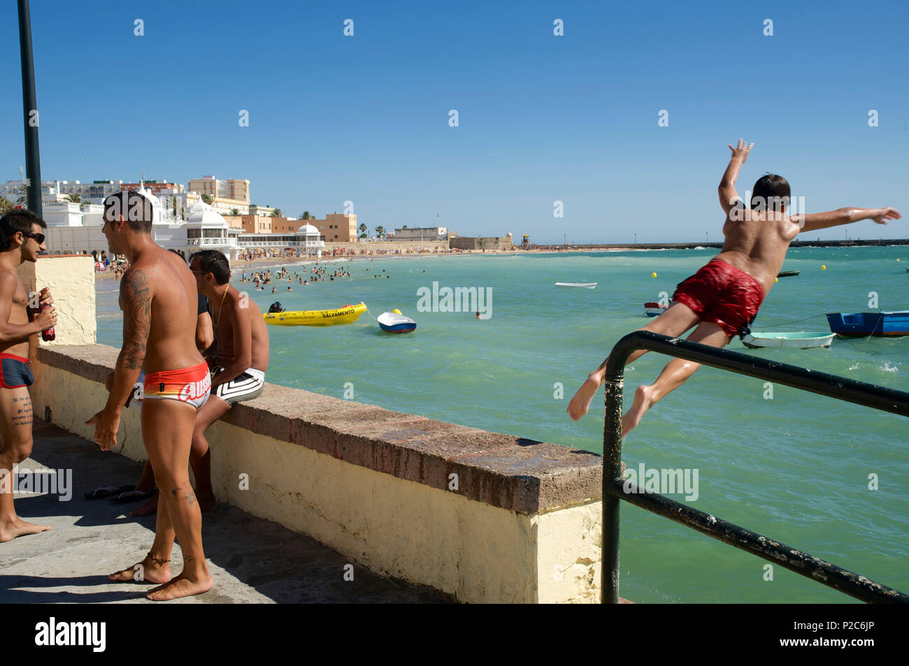 3 men on the beach and one boy jumping from the harbour wall into the sea next to the Castillo de Santa Catalina, Cadiz, Andalus Stock Photo