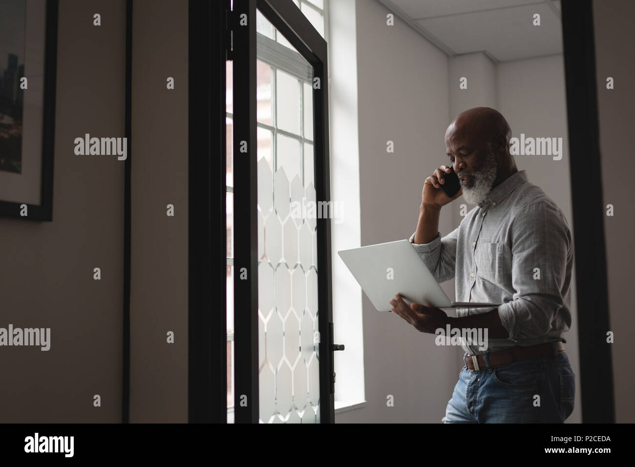 Senior graphic designer using laptop while talking on mobile phone Stock Photo