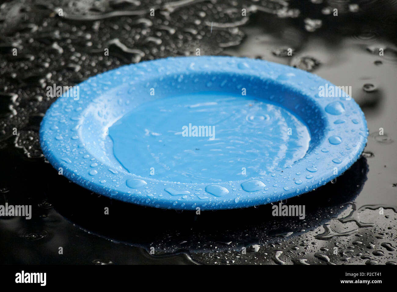 blue plastic plate sitting on black garden table filling up with water due to heavy rain on typical british summer day Stock Photo