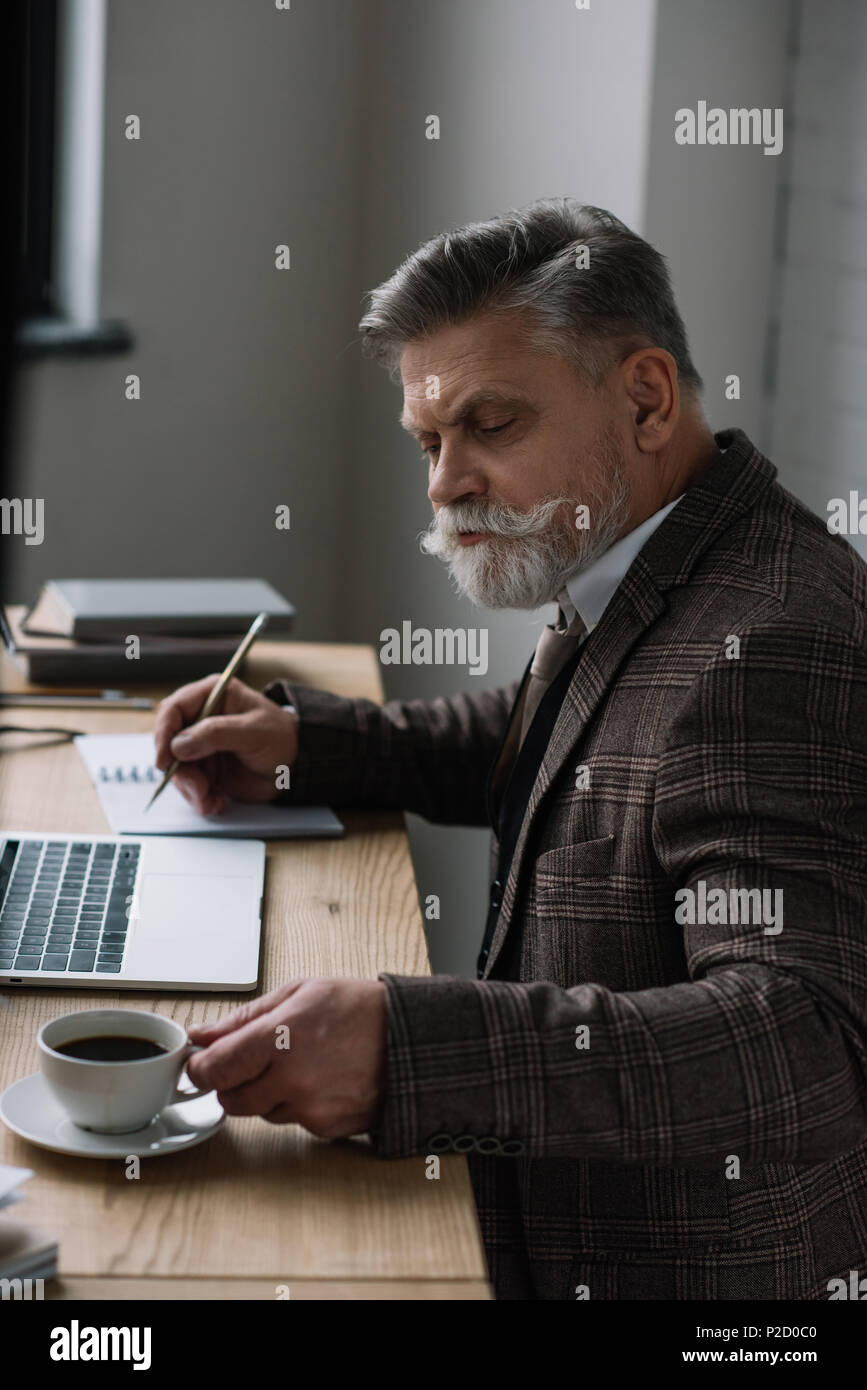serious senior writer working with laptop and making notes in notebook ...