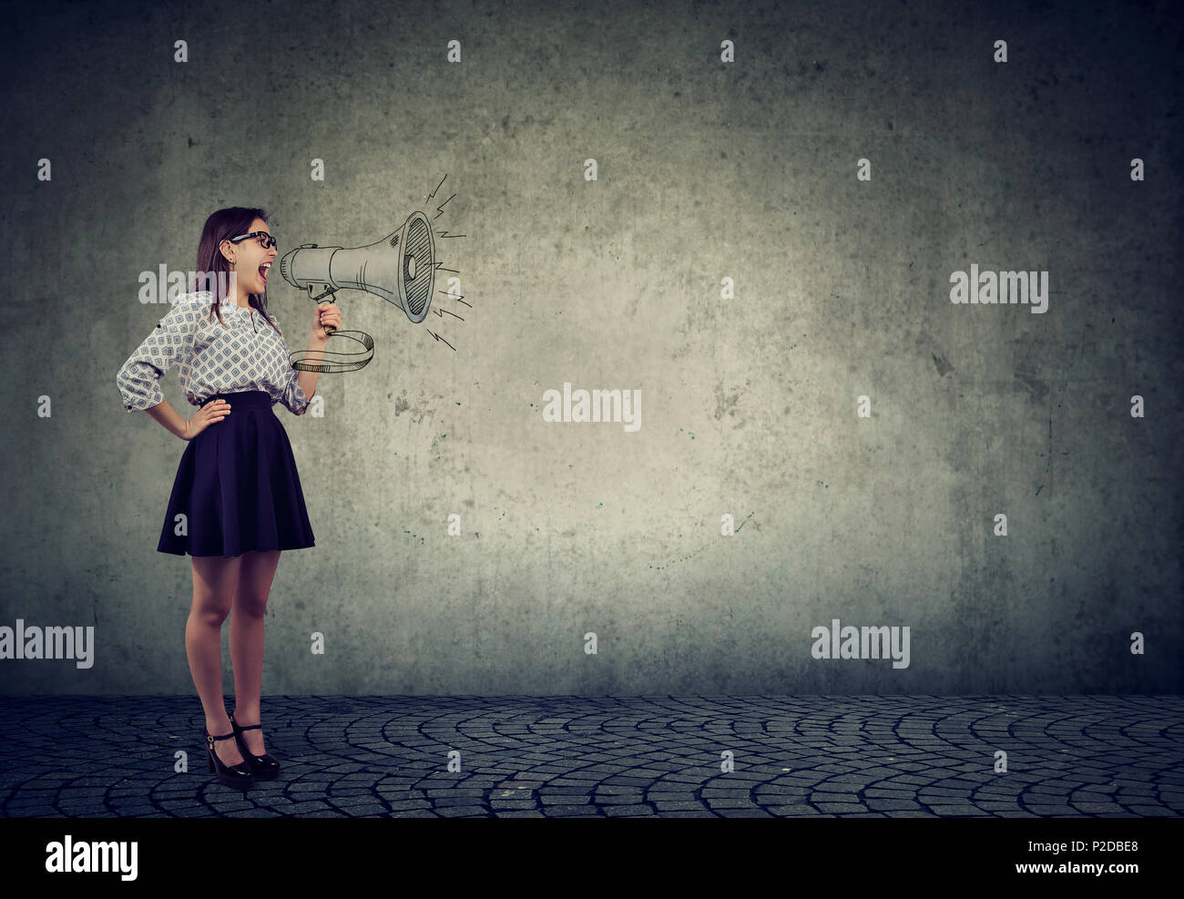Side view of young girl screaming and announcing news with megaphone against gray wall. Stock Photo