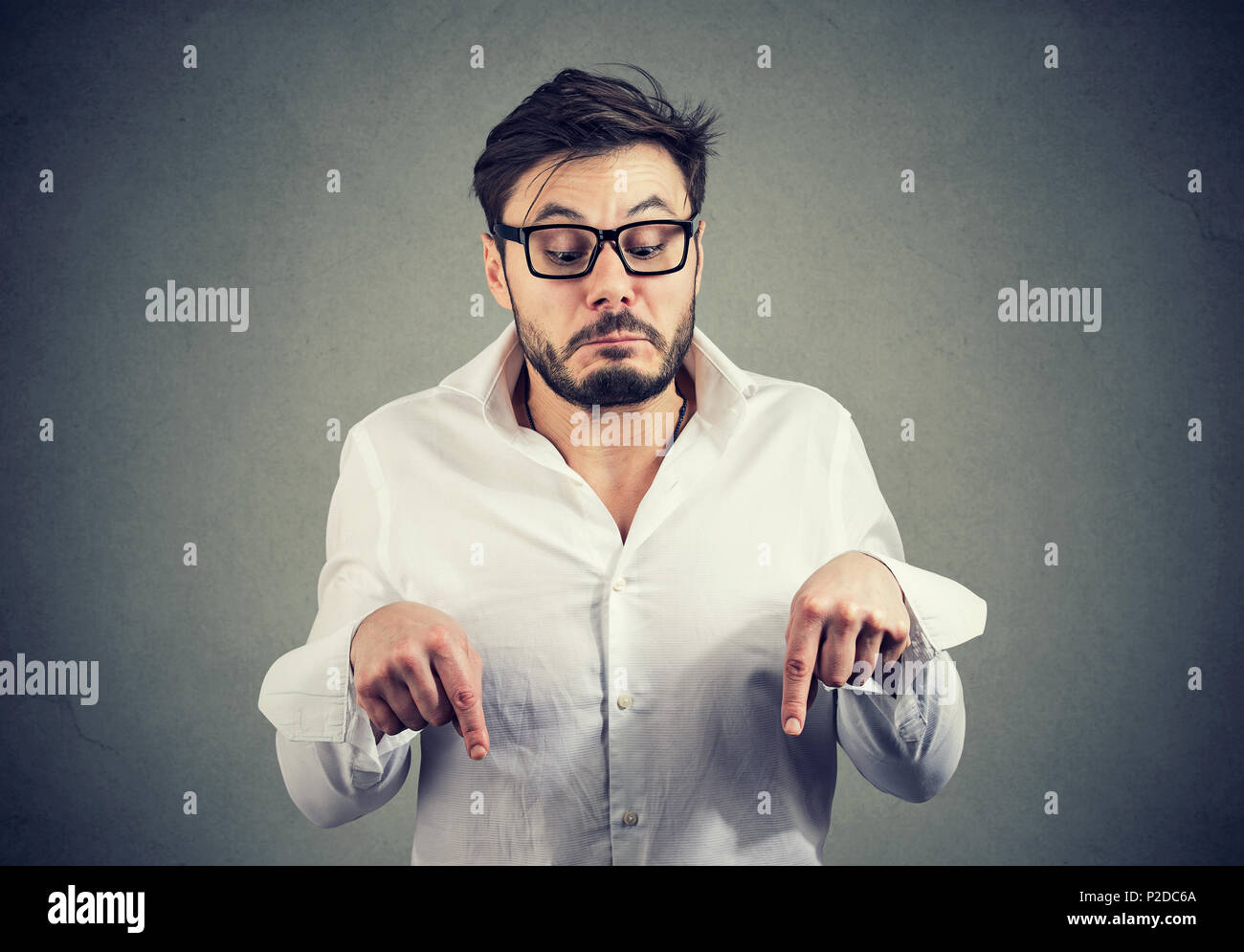 Young bearded man looking funny while directing down with fingers on gray background Stock Photo