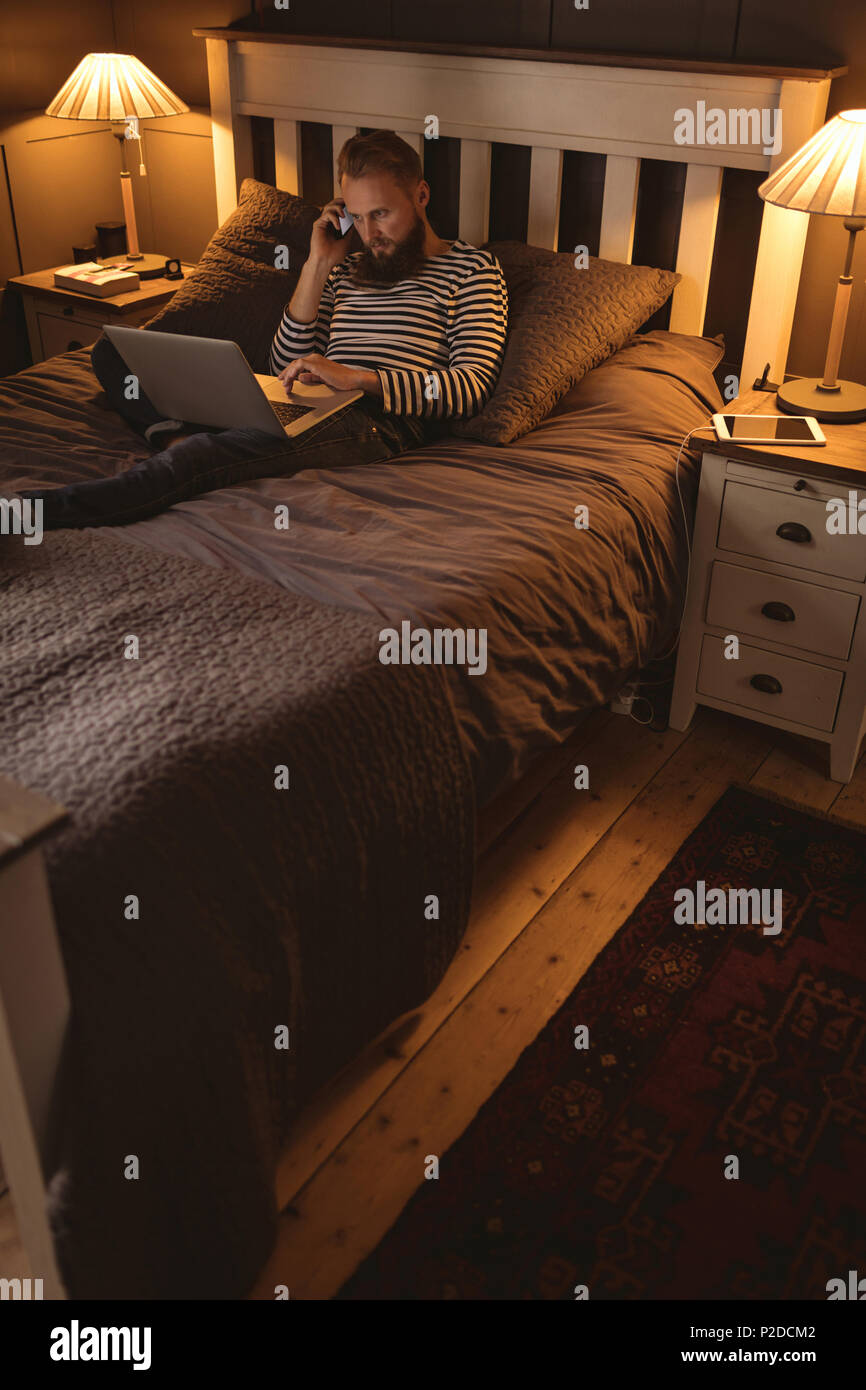 Man lying on bed talking on phone while using laptop Stock Photo