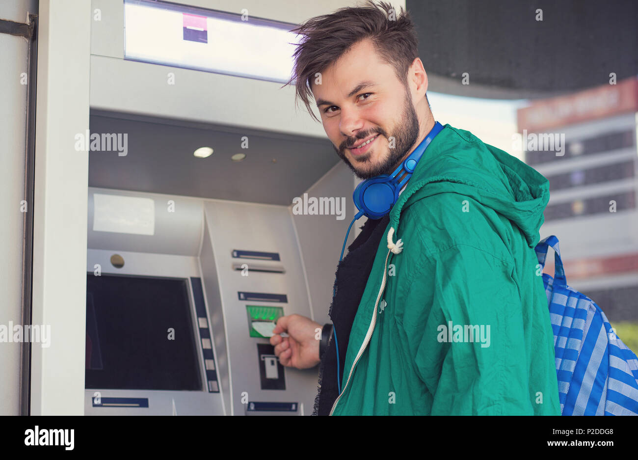 Young hipster guy in headphones smiling at camera while inserting credit card in ATM machine getting cash money Stock Photo