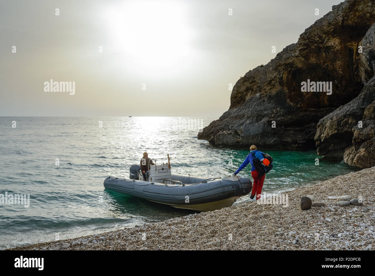 Young woman with trekking gear embarking an inflatable boat on the pebble beach of the bay Cala Sisine that will bring her back Stock Photo
