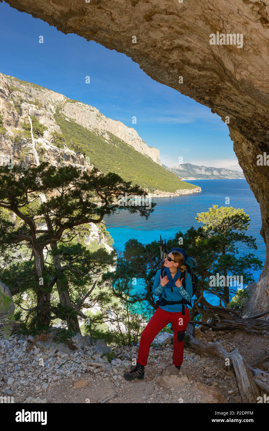 A young woman with trekking gear hikes through the rock arch Arcu su Feilau at the mountainous coast above the sea, Golfo di Oro Stock Photo