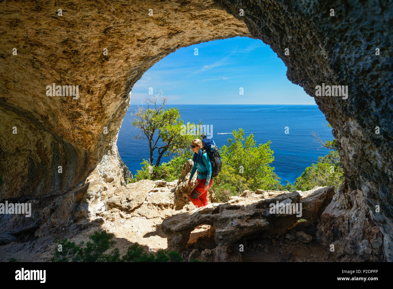 A young woman with trekking gear hiking through the rock arch Arcu su Feilau at the mountainous coast above the sea, Golfo di Or Stock Photo