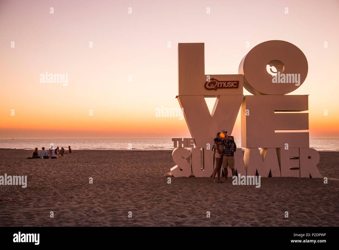 Couple taking a selfie photograph with smartphone in front of Love sculpture on the beach at sunset, Ostend, Flanders, Flemish R Stock Photo