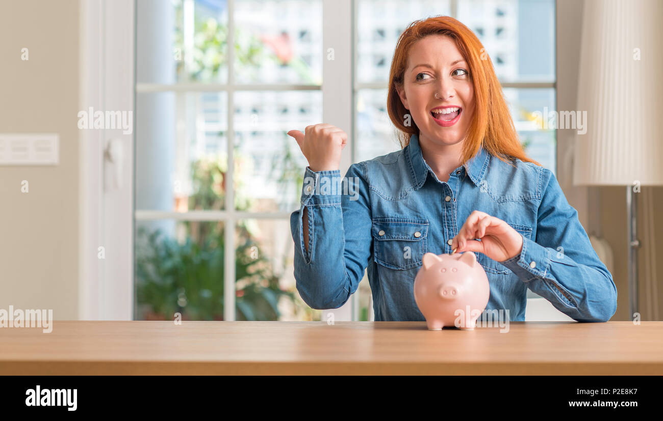 Redhead woman saves money in piggy bank at home pointing with hand and ...