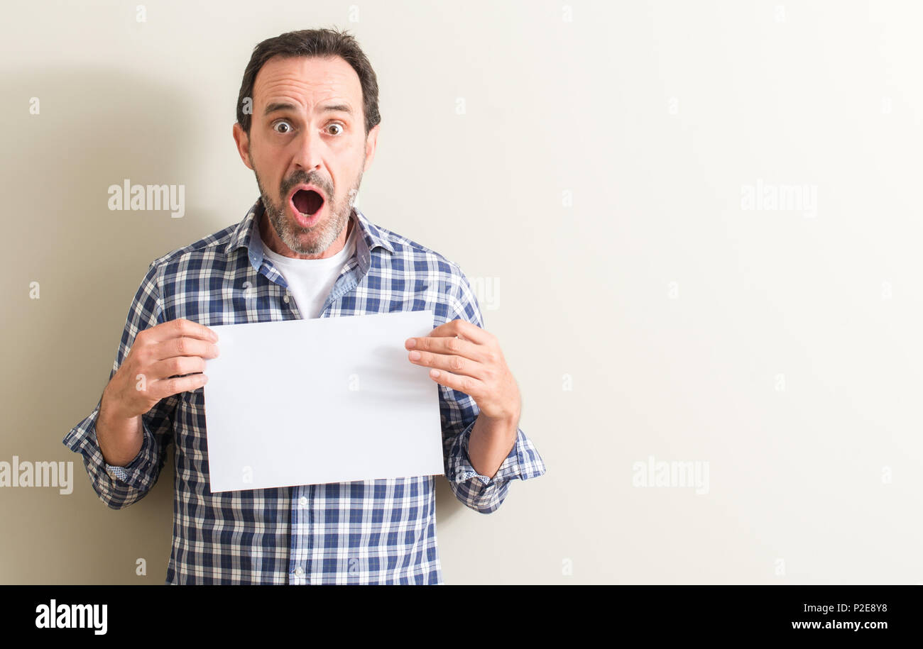 Senior man holding blank paper sheet scared in shock with a surprise