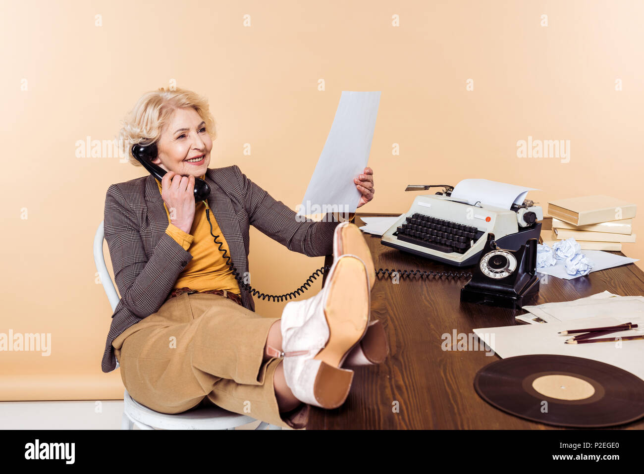 smiling senior woman with legs on table talking on rotary phone and looking at pape Stock Photo